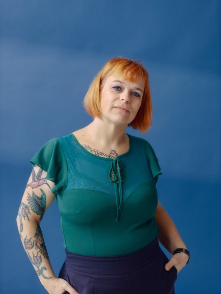 Marjolein draagt een turkooisblauw shirt en heeft tattoos op haar armen. 