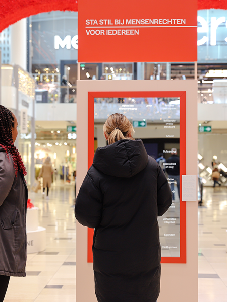 De Mensenrechtenspiegel staat in een passage van winkelcentrum Hoog Catharijne. Een vrouw kijkt in een spiegel, terwijl een andere vrouw toekijkt. 