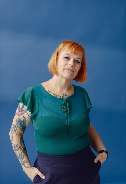 Marjolein draagt een turkooisblauw shirt en heeft tattoos op haar armen. 